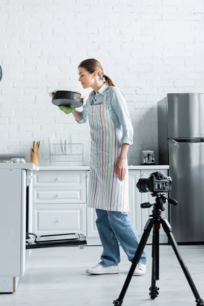 Blogueiro culinário satisfeito cheirando forma de cozimento perto de câmera digital na cozinha — Fotografia de Stock