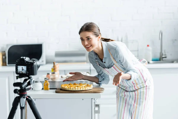 Счастливый кулинарный блогер указывая руками на вкусный пирог возле размытой цифровой камеры — стоковое фото