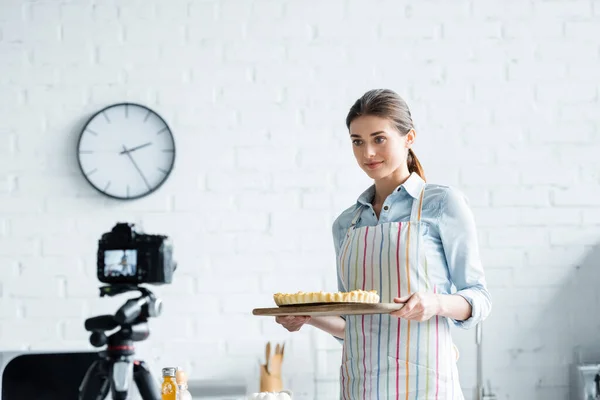 Joven mujer en delantal mirando borrosa cámara digital mientras sostiene sabroso pastel - foto de stock