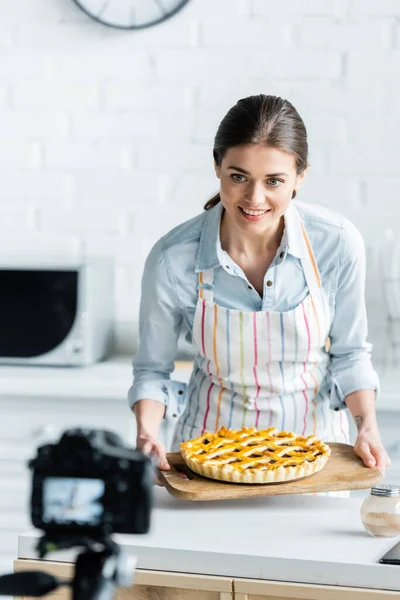 Blurred digital camera near happy culinary blogger holding tasty pie — Stock Photo