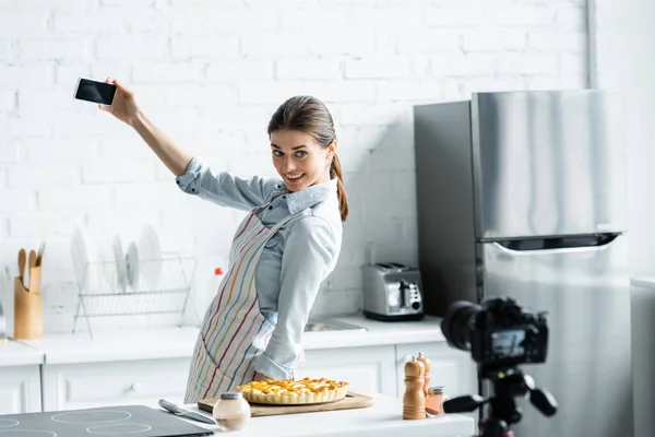 Blogueiro culinário alegre tomando selfie com torta saborosa e câmera digital na cozinha — Fotografia de Stock