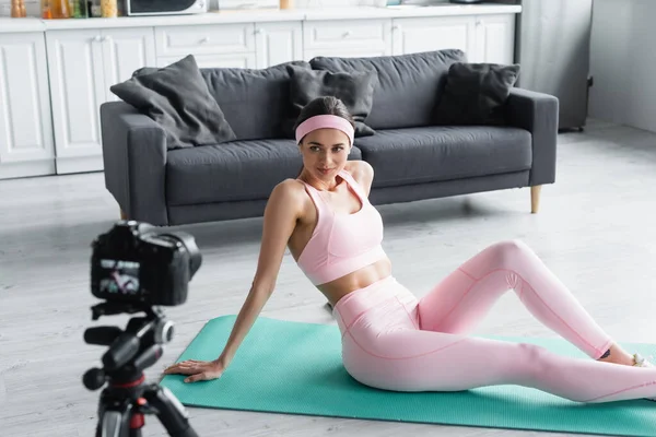 Mujer joven en ropa deportiva sentada en la colchoneta de fitness cerca de la cámara digital borrosa - foto de stock