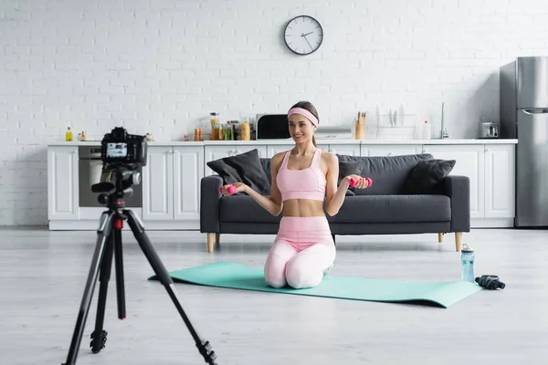 Smiling sports blogger with dumbbells talking while recording lesson on digital camera — Stock Photo