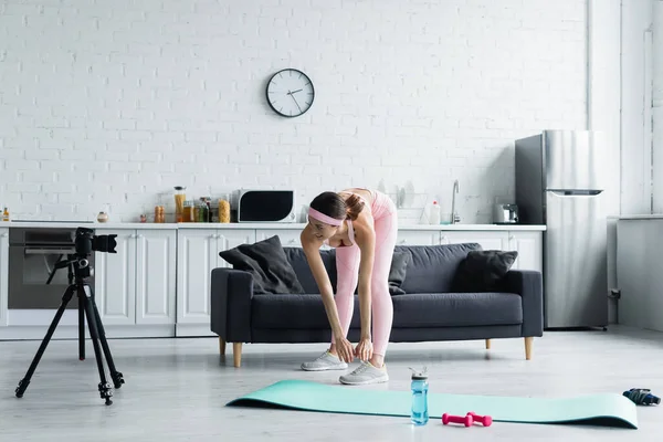 Young sports blogger doing forward lunges near digital camera on tripod at home — Stock Photo