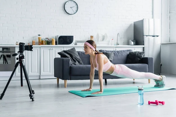 Young sports blogger exercising in plank pose near digital camera in kitchen — Stock Photo