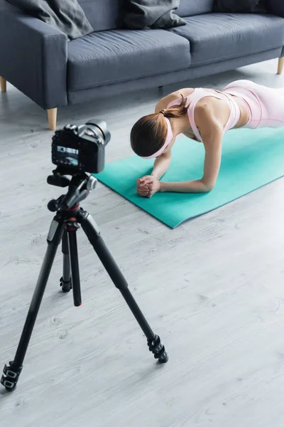 Appareil photo numérique sur trépied près d'entraînement sportif en pose de planche sur tapis de fitness — Photo de stock