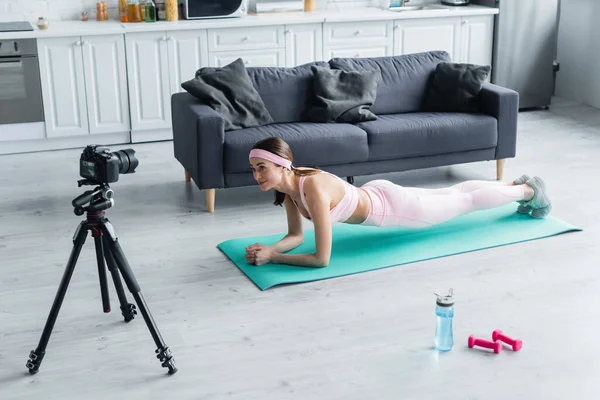 Bastante bloguero deportivo mirando a la cámara digital mientras hace ejercicio en la pose tablón en la alfombra de fitness - foto de stock