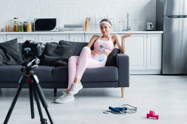 Junge Sportlerin mit Sportflasche auf Sofa vor verschwommener Digitalkamera — Stockfoto