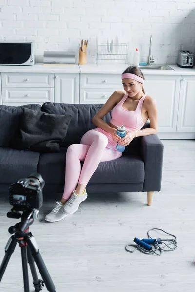Donna con bottiglia sportiva seduta sul divano vicino alla fotocamera digitale su treppiede — Foto stock