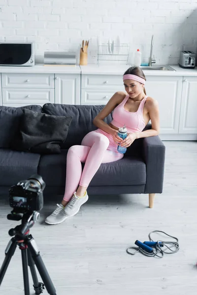 Frau mit Sportflasche sitzt auf Sofa neben Digitalkamera auf Stativ — Stock Photo