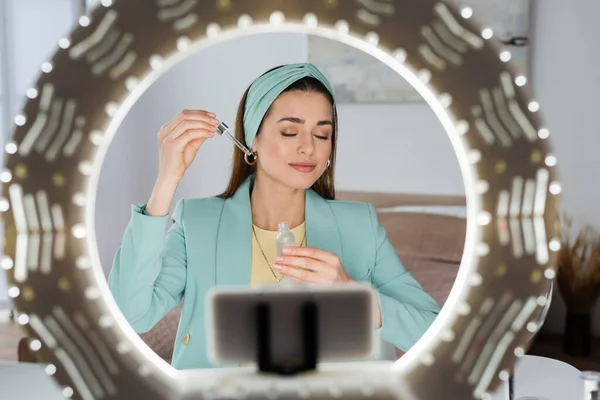 Teléfono inteligente borroso en el titular con luz de anillo cerca de belleza blogger aplicar suero facial — Stock Photo