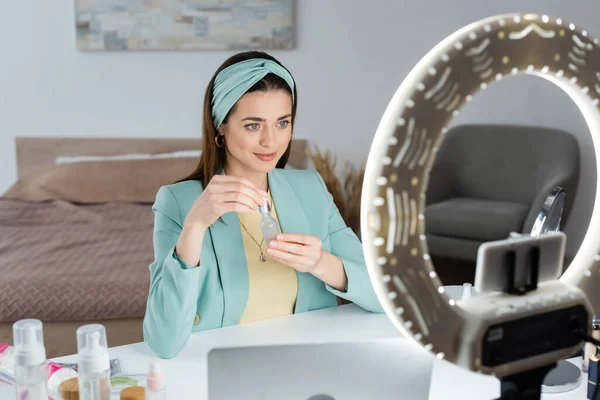 Mujer elegante que muestra suero facial cerca de ordenador portátil borroso y teléfono inteligente en la lámpara de anillo - foto de stock