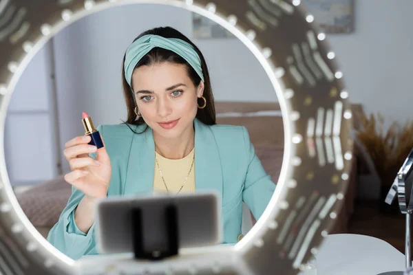 Young beauty blogger proposing lipstick near phone holder with circle lamp — Stock Photo