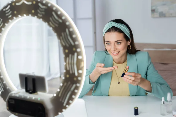 Smiling woman pointing at lipstick near smartphone in holder with circle lamp — Stock Photo