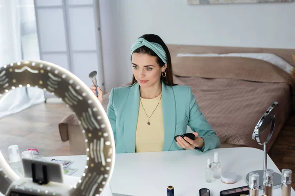 Beauty-Bloggerin hält Kosmetikpinsel und Gesichtspuder in der Nähe eines Telefonhalters mit Ringlampe — Stockfoto