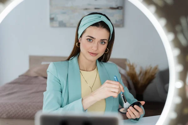 Young beauty blogger holding face powder and cosmetic brush near blurred phone holder with ring lamp — Stock Photo