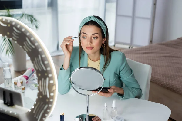 Bella donna applicando ombretti vicino specchio e smartphone in titolare con luce anulare — Foto stock