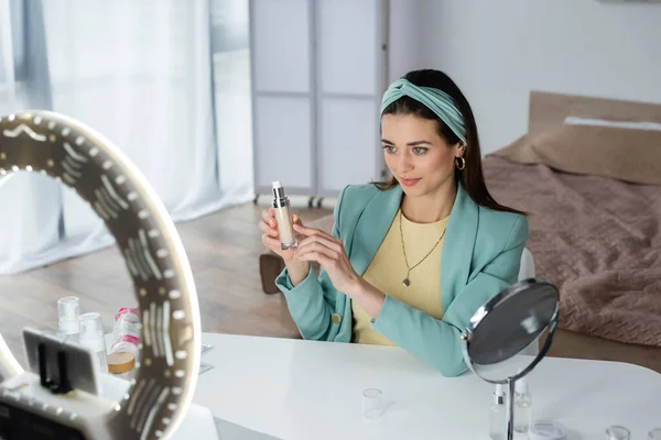 Femme élégante pointant vers le fond de teint près du support de téléphone flou avec lampe annulaire — Photo de stock