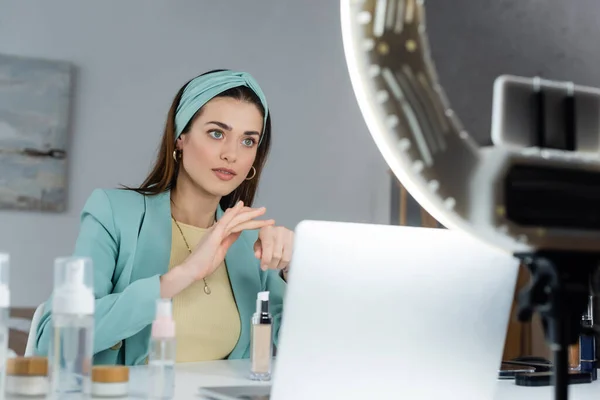 Pretty woman applying face foundation on hand near blurred laptop and phone holder — Stock Photo