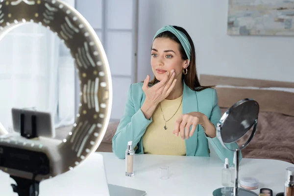 Stylish beauty blogger applying face foundation near blurred laptop and phone holder with ring lamp — Stock Photo