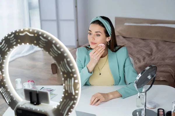 Beauty blogger wiping face with cosmetic sponge near phone holder with ring light — Stock Photo