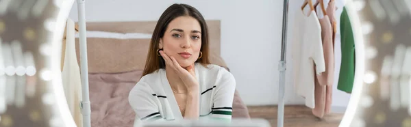 Bastante blogger de moda mirando a la cámara cerca de perchas con ropa y lámpara de círculo borrosa, pancarta — Stock Photo