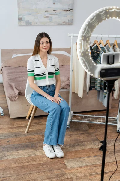 Pretty fashion blogger sitting near hangers with clothes and phone holder with ring lamp — Stock Photo