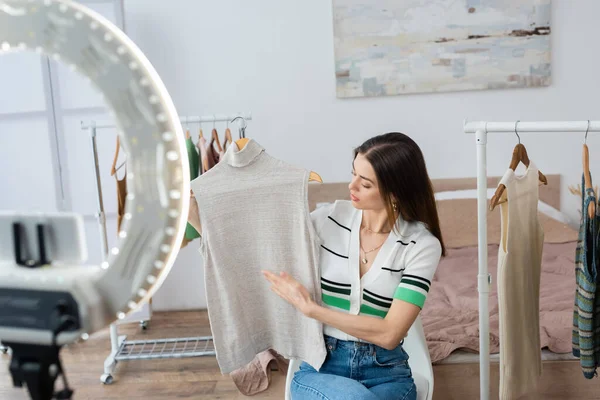 Blogueiro de moda apontando para o colete perto do suporte de telefone borrado com lâmpada de anel — Fotografia de Stock