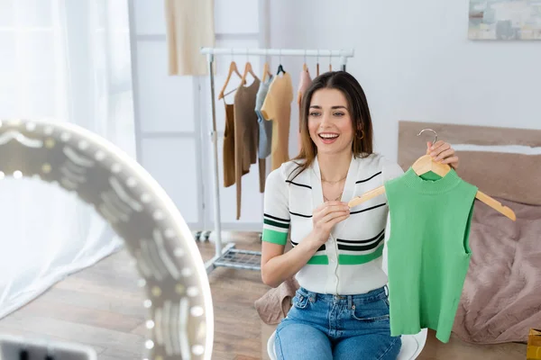 Cheerful fashion blogger holding hanger with vest near blurred ring lamp — Stock Photo