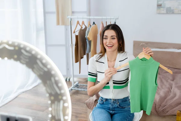 Alegre blogger de moda sosteniendo percha con chaleco cerca de la lámpara de anillo borrosa - foto de stock