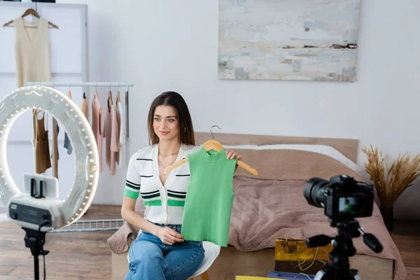 Pleased fashion blogger showing vest on hanger near blurred digital camera and phone holder — Stock Photo