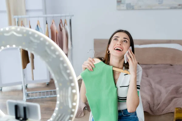 Excited fashion blogger talking on smartphone while holding vest near phone holder with ring lamp — Stock Photo