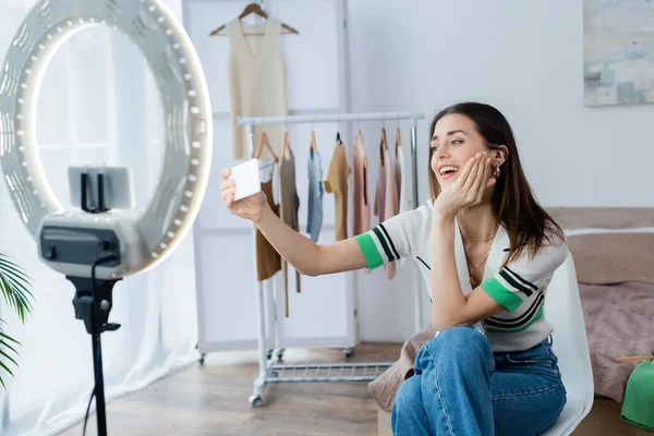 Blogger de moda feliz tomando selfie cerca de perchas con ropa y soporte para teléfono con lámpara de anillo - foto de stock