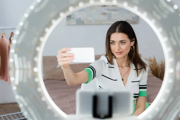 Titular de teléfono borroso con lámpara de anillo cerca de blogger de moda tomando selfie en el teléfono inteligente - foto de stock