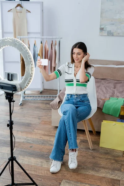 Blogger de moda bonita y positiva tomando selfie cerca de la ropa y el titular del teléfono con lámpara de círculo - foto de stock