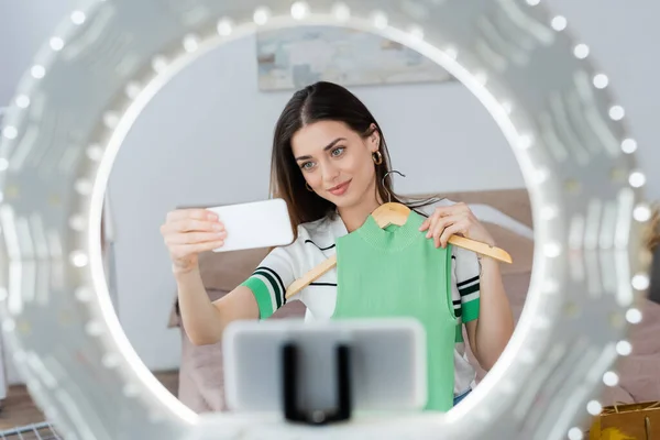 Lächelnde Mode-Bloggerin macht Selfie mit Weste auf Kleiderbügel neben verschwommenem Handyhalter mit Ringlicht — Stockfoto
