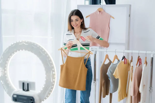 Joyful fashion blogger holding tag with sale lettering near clothes and phone holder — Stock Photo