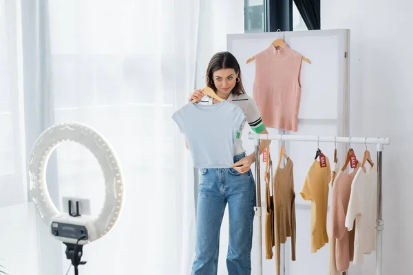 Bastante blogger de moda mostrando camiseta cerca del titular del teléfono con lámpara de círculo - foto de stock