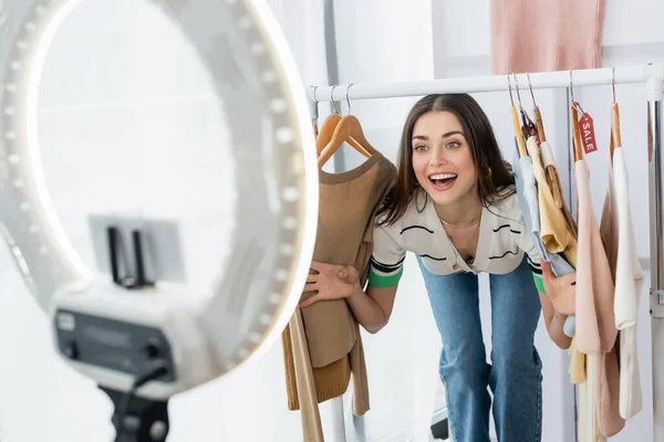 Allegro fashion blogger vicino appendini con vestiti e porta telefono sfocato con luce anulare — Foto stock