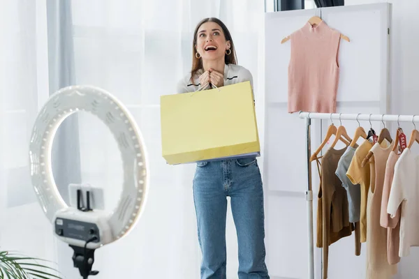 Mujer sorprendida con bolsa de compras cerca de la ropa y titular del teléfono con lámpara de círculo - foto de stock