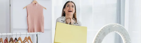 Blogueur de mode étonné tenant sac à provisions près des vêtements et lampe annulaire, bannière — Photo de stock