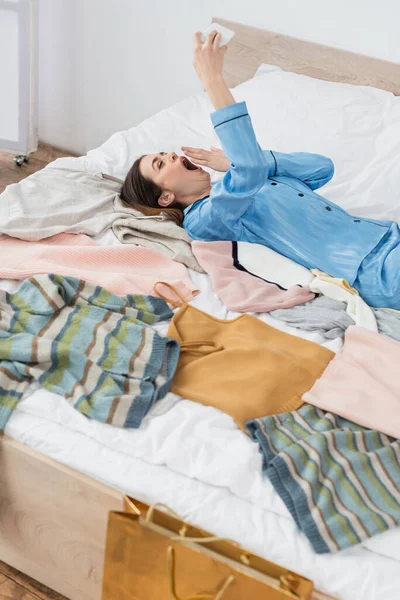 Mujer cansada bostezando durante la videollamada cerca de un montón de ropa en la cama - foto de stock