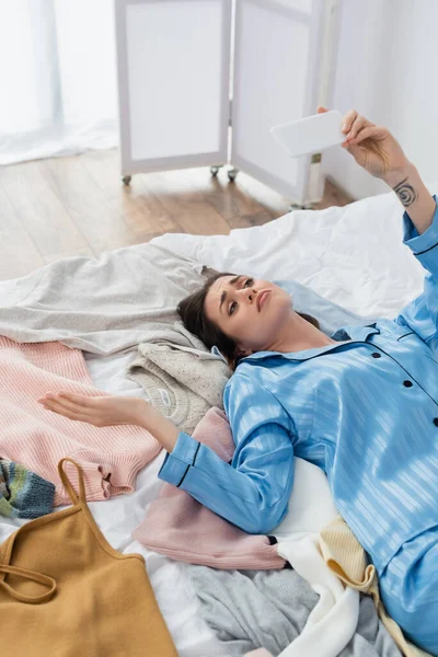 Mulher descontente no elegante pijama gesto durante chamada de vídeo no smartphone perto de roupas diferentes — Fotografia de Stock
