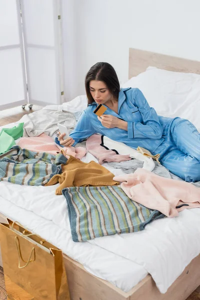 Frau im stylischen Pyjama mit Handy in der Nähe von viel Kleidung im Schlafzimmer — Stockfoto