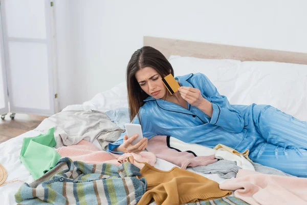 Mujer molesta en pijama elegante mirando el teléfono inteligente mientras sostiene la tarjeta de crédito cerca de un montón de ropa - foto de stock