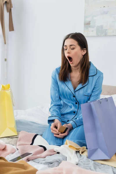 Sleepy woman in stylish pajama yawning while sitting with credit card near shopping bags and clothes — Stock Photo