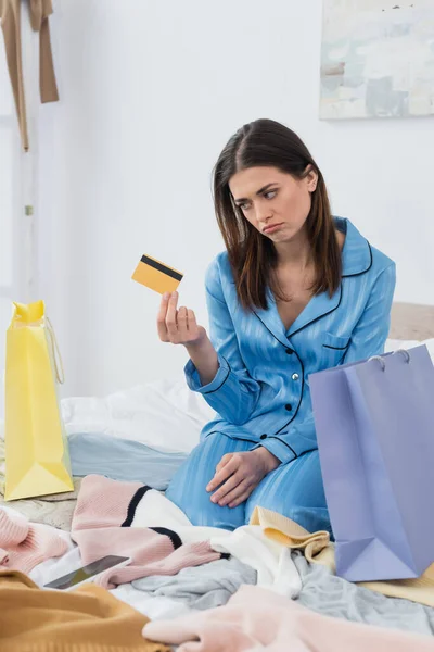 Mujer ofendida con tarjeta de crédito sentado cerca de bolsas de compras y ropa en el dormitorio - foto de stock