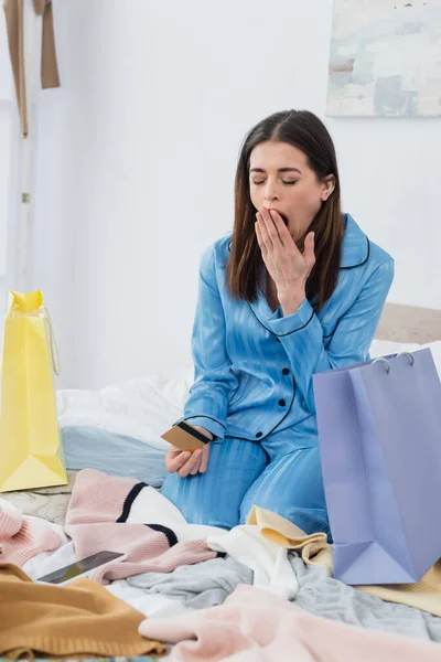 Femme fatiguée en pyjama élégant bâillant près des sacs à provisions et assortiment de vêtements sur le lit — Photo de stock