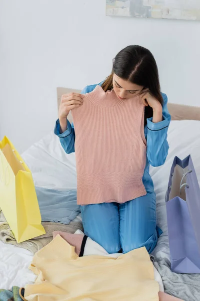 Woman in silk pajama holding vest while sitting on bed near shopping bags and clothing — Stock Photo