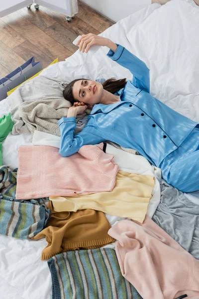 Smiling blogger in silk pajama using smartphone while lying near assortment of clothes on bed — Stock Photo